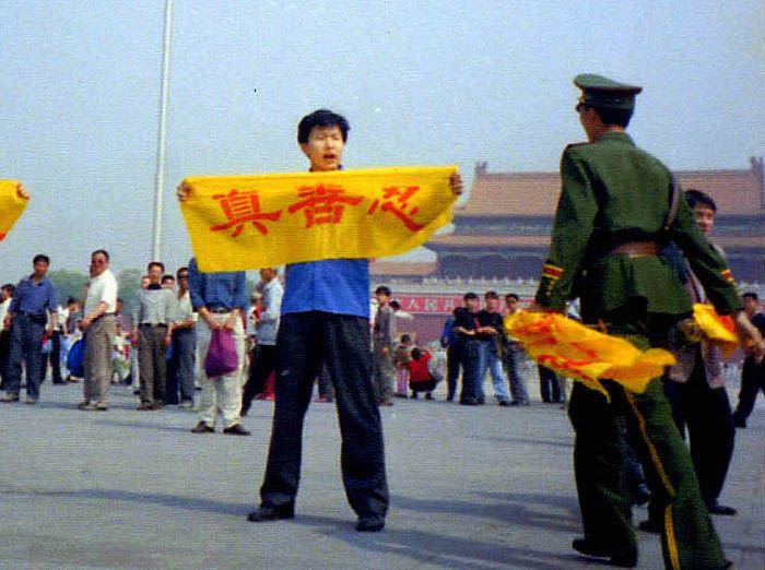 Китайски полицай се приближава до практикуващ Фалун Гонг на площад "Тиенанмън" в Пекин, докато той държи плакат с китайските йероглифи за "истинност, състрадание и търпение" - основните принципи на Фалун Гонг (С любезното съдействие на Minghui.org)