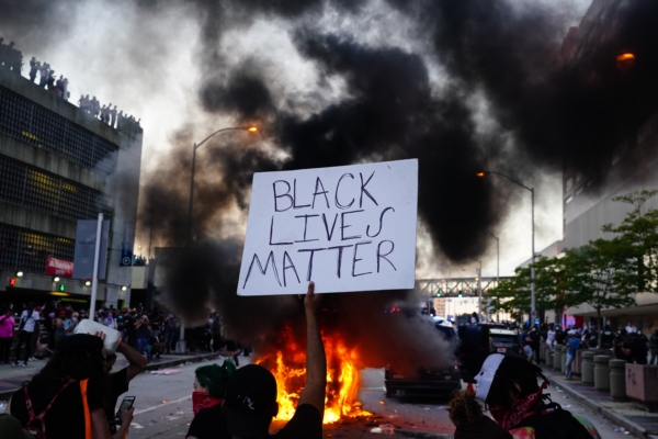 Мъж държи знак Black Lives Matter, докато пред него гори полицейска кола по време на протест заради смъртта на Джордж Флойд, пред CNN Center в Атланта на 29 май 2020 г. (Elijah Nouvelage/Getty Images)