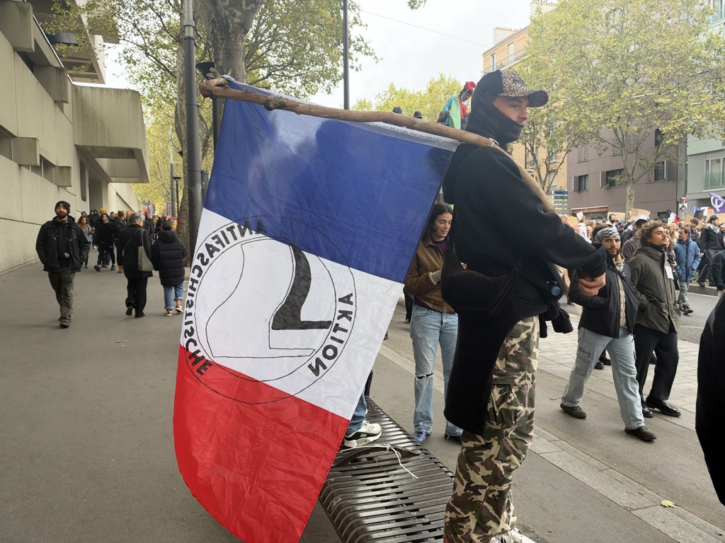 Френските протести отново прераснаха в насилие, но масираното полицейско присъствие ограничи грабежите 22 Маскиран протестиращ носи френско знаме със символ на Антифа по време на демонстрация в Париж на 24 октомври 2026 г., на фона на крайнолеви протести в цяла Франция заради нарастващите разходи за живот и съкращенията на финансирането за образование. (Etienne Fauchaire/The Epoch Times)